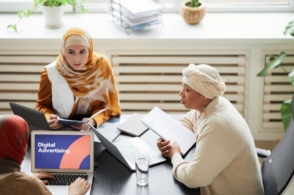 pexels photo 8154578 8154578 Three women in hijabs collaborating on digital advertising strategies around a table with laptops.